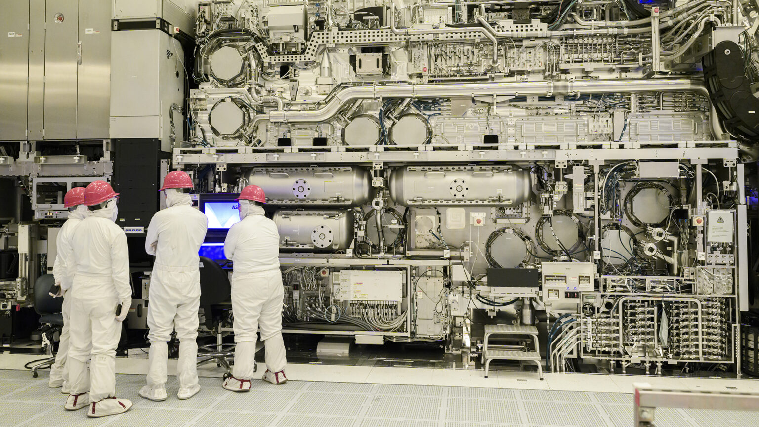 Technicians in cleanroom suits and red hard hats near large semiconductor manufacturing equipment in a sterile fabrication facility